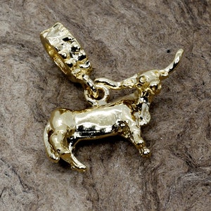 May include: A gold-colored charm in the shape of a bull with its head turned to the side. The bull has a textured surface and a ring for attaching it to a bracelet or necklace.