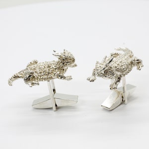 May include: A pair of silver cufflinks featuring a detailed, textured design of a horned lizard. The cufflinks are attached to a silver base.
