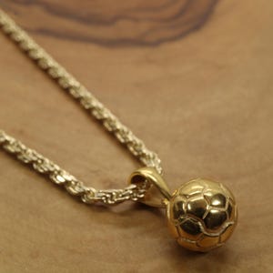 Puede incluir: Un collar con colgante de balón de fútbol dorado. El colgante es un balón de fútbol en miniatura detallado, suspendido de una cadena dorada a juego. El collar se muestra sobre un fondo de madera cálido.