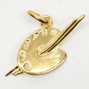 Puede incluir: Un dije dorado con forma de paleta de artista con un pincel. La paleta tiene varios huecos para pintura y un lazo para sujetarlo a un collar o pulsera. El dije está hecho de un metal brillante y reflectante.