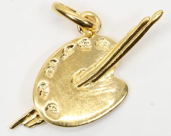 May include: A gold-coloured charm depicting an artist's palette and paintbrush. The palette features several paint wells and a loop for attachment. The charm is made of a shiny, reflective metal, suitable for a necklace or bracelet.