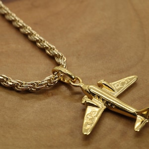 May include: A gold-colored airplane pendant necklace. The pendant is a detailed miniature airplane, suspended from a thick, textured gold chain. The necklace is displayed on a brown surface.