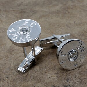 Może przedstawiać: Para srebrnych spinek do mankietów w kształcie łusek od strzelby z małym diamentem w środku każdej. Spinki do mankietów są grawerowane tekstem "FC ONE" i "FC ONE".