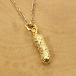 Peut inclure: Un collier doré avec un pendentif en forme de cornichon. Le pendentif a une surface texturée et est suspendu à une fine chaîne dorée. Le collier est présenté sur un fond neutre.