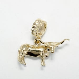 May include: A gold-colored charm in the shape of a longhorn steer. The charm has a detailed design, including the steer's body, legs, head, and curved horns. The charm has a loop at the top for attaching to a bracelet or necklace.