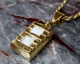 Puede incluir: Collar de cadena de oro con un colgante con forma de ladrillo con dos ventanas rectangulares blancas.