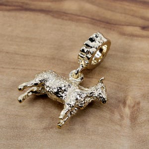 Könnte beinhalten: Ein goldfarbener Anhänger in Form eines laufenden Lamms. Der Anhänger hat eine strukturierte Oberfläche und ist an einem ringförmigen Bügel aufgehängt. Das Lamm ist detailliert mit sichtbaren Beinen, Körper und Kopf.