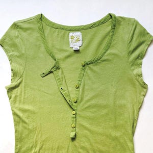 May include: A light green short-sleeved Henley shirt with a button-down placket. The shirt has a scoop neckline and a small tag near the collar. The fabric appears to be a soft, lightweight material.