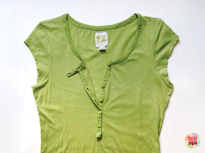 May include: A light green short-sleeved Henley shirt with a button-down placket. The shirt has a scoop neckline and a small tag near the collar. The fabric appears to be a soft, lightweight material.