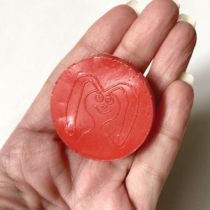 May include: A red, circular toy with a rabbit design. The toy is held in a hand and features a smiling rabbit face with long ears. The rabbit design is embossed on the surface of the toy.