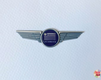 May include: A silver-toned US Airways pilot wing pin. The pin features a circular emblem with the US flag and the words "US AIRWAYS". The wings are detailed with multiple feather-like ridges.