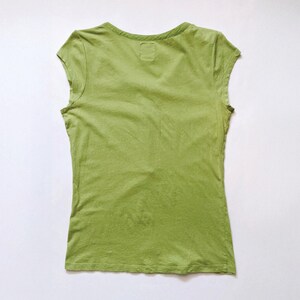 May include: A sleeveless, light green t-shirt with a V-neck. The shirt is made of a soft, slightly textured fabric. The back of the shirt is visible, and it is laid flat on a white surface.