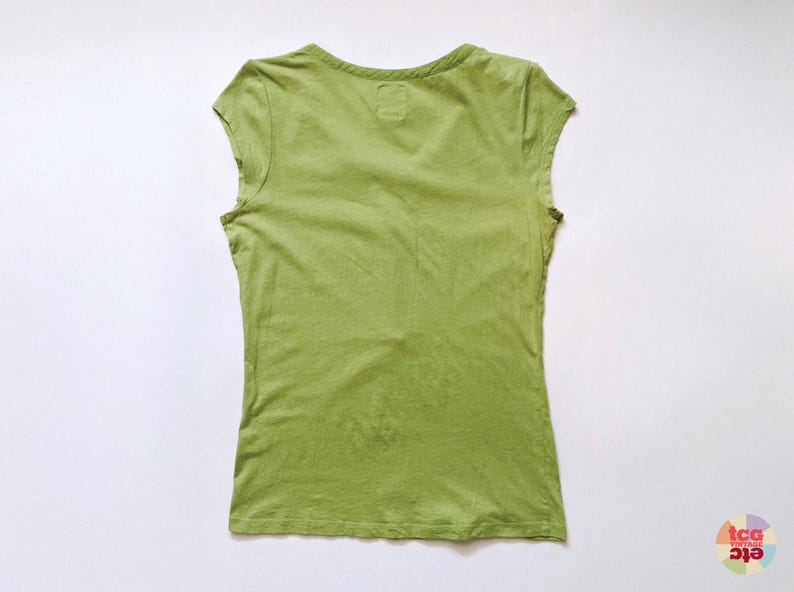 May include: A sleeveless, light green t-shirt with a V-neck. The shirt is made of a soft, slightly textured fabric. The back of the shirt is visible, and it is laid flat on a white surface.
