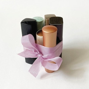 May include: A collection of lipsticks in various colors, including black, gold, and beige, tied together with a light purple ribbon. The lipsticks are arranged in a bundle, creating a visually appealing presentation.