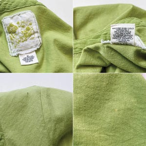 May include: Close-up of a light green shirt. The shirt has a tag with a floral design and another tag with washing instructions. The fabric appears to be a soft cotton material. The shirt is a medium size.