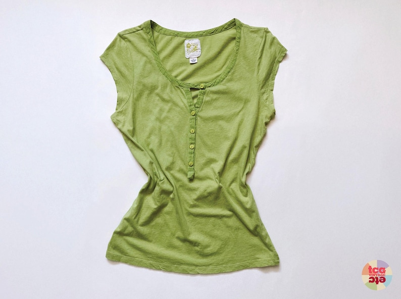 May include: A light green, short-sleeved shirt with a scoop neck and a button-down placket. The shirt has a slightly faded appearance and a small tag near the neckline. The shirt is made of a soft, lightweight fabric.