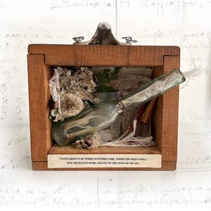 Puede incluir: Una caja de madera con una botella de vidrio en su interior. La botella está llena de una sustancia blanca y un trozo de papel. La caja está decorada con flores secas y otros elementos naturales. El texto en el papel dice: "He pedido estar donde no llegan las tormentas, donde la ola verde está en los puertos mudos, y fuera del balanceo del mar."