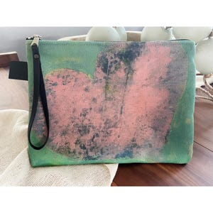 Könnte beinhalten: Eine grüne Clutch aus Canvas mit einem abstrakten Design in Pink und Schwarz. Die Tasche hat einen schwarzen Reißverschluss und einen schwarzen Riemen.