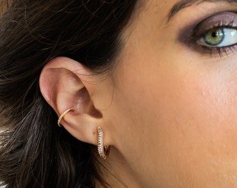 Puede incluir: Un ear cuff dorado y un pequeño pendiente de aro dorado a juego con piedras transparentes incrustadas. El ear cuff se coloca en el cartílago superior, mientras que el aro se lleva en el lóbulo, ambos con un acabado metálico pulido.