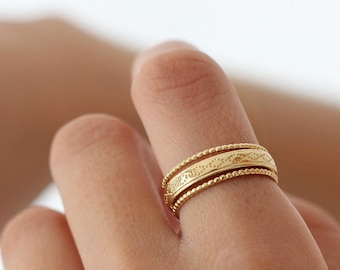 May include: A gold ring with a detailed design is worn on a finger. The ring features a central band with an etched pattern, bordered by beaded edges. The ring's warm gold color is prominent against the skin.