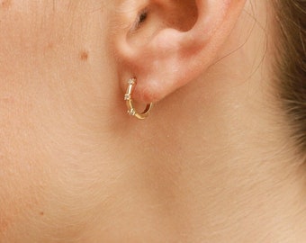 Puede incluir: Primer plano de la oreja de una persona con dos pendientes de oro. El pendiente del lóbulo inferior es un aro pequeño, y el pendiente del lóbulo superior es una fila de pequeños diamantes.