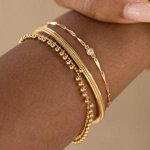 Peut inclure: Un ensemble de trois bracelets en or, l'un avec une chaîne délicate, l'autre avec une frange de perles et le dernier avec une bande texturée. Les bracelets sont empilés sur le poignet d'une personne.