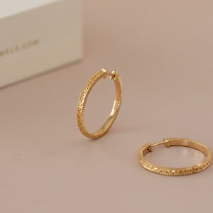 May include: A pair of gold hoop earrings with a textured design. The earrings are on a light pink background.