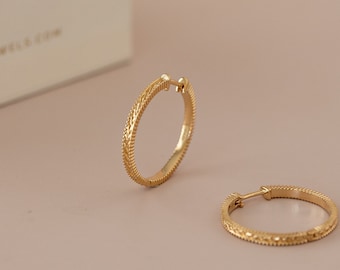 Peut inclure: Une paire de boucles d'oreilles créoles en or avec un motif texturé. Les boucles d'oreilles sont sur un fond rose clair.