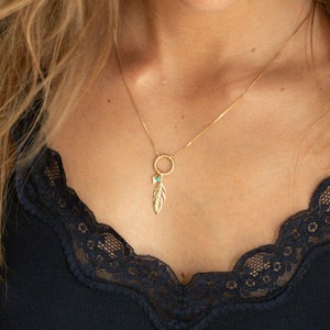 Può includere: Una collana d'oro con un ciondolo a forma di piuma e una piccola pietra turchese. La collana è indossata da una persona con i capelli lunghi biondi e una canotta nera con bordino in pizzo.