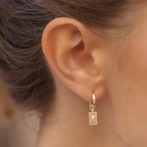 Könnte beinhalten: Goldene Creolen mit einem kleinen rechteckigen Anhänger. Der Anhänger hat einen kleinen Diamanten in der Mitte.