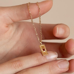 Puede incluir: Un collar de cadena de oro con un pequeño colgante rectangular que presenta una piedra preciosa roja en el centro. El colgante tiene un diseño detallado e intrincado.