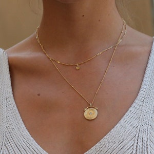 Puede incluir: Una mujer lleva un top blanco de punto de tanque con un collar de cadena dorada que presenta un pequeño dije de estrella y un dije circular más grande con un diseño detallado.