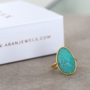 May include: A gold ring featuring a large, oval-shaped green gemstone. The ring is displayed on a grey surface.