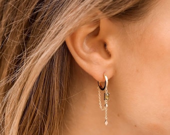 May include: Gold hoop earrings with a delicate chain and a small green gemstone dangling from the bottom.