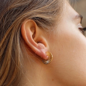 Puede incluir: Pendientes de aro dorados con un diseño de sol. Los pendientes son usados por una persona con cabello largo y castaño.