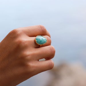 May include: A gold ring featuring a large, oval-shaped, light blue gemstone. The ring is set against a blurred backdrop of water and rocks.