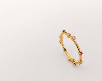 May include: A delicate gold ring with colorful gemstones. The ring features a series of small, evenly spaced gemstones in shades of pink, red, and blue. The ring is set against a white background.