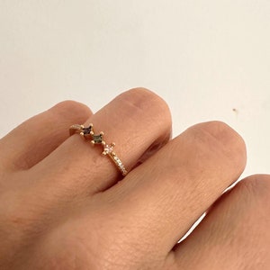 May include: A gold ring with three square-shaped gemstones. The gemstones are a dark green, a dark purple, and a light pink. The ring is set with small clear stones.
