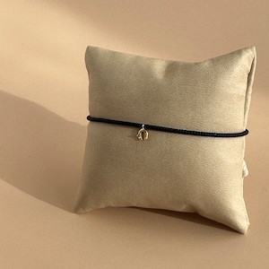 May include: A black string bracelet with a gold charm in the shape of an arch. The bracelet is displayed on a light beige pillow.