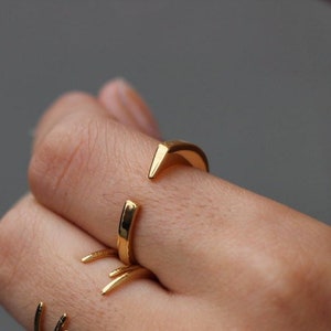 May include: A hand wearing four gold rings. The rings are all different styles, with some being simple bands and others being more intricate.