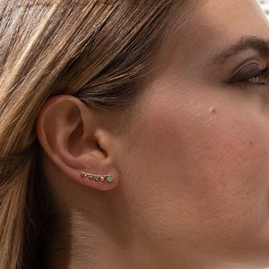 Może przedstawiać: Kobieta z długimi blond włosami nosi parę złotych kolczyków z małymi kamieniami szlachetnymi. Kolczyki są proste w designie i idealne do codziennego noszenia.