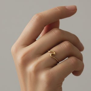 May include: A gold-colored ring with a wave design. The ring is thin and delicate, with a smooth, polished finish. The wave shape is the focal point, creating a minimalist and elegant aesthetic. The ring is worn on a finger.