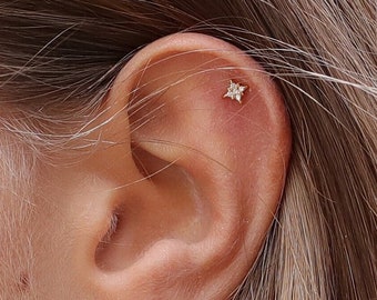 Puede incluir: Una oreja con tres pendientes de estrella de oro. Dos de los pendientes son pequeños y uno es un aro con un colgante de estrella.