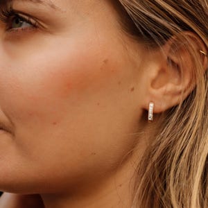 Könnte beinhalten: Nahaufnahme eines Ohrs mit einem kleinen, goldenen Creolen-Ohrring. Der Ohrring ist mit kleinen, klaren Edelsteinen besetzt. Der Hautton ist hell, und das Haar ist hellbraun und wellig.