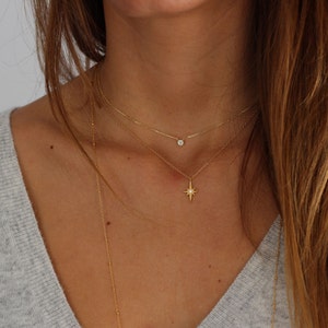 Puede incluir: Un collar de cadena de oro con un peque&ntilde;o colgante de diamante y un colgante de estrella m&aacute;s grande. El collar est&aacute; superpuesto en el cuello de una modelo.