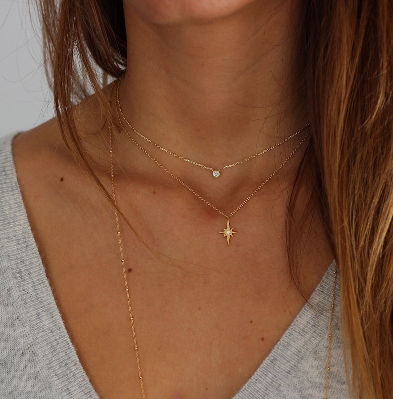 Puede incluir: Un collar de cadena de oro con un peque&ntilde;o colgante de diamante y un colgante de estrella m&aacute;s grande. El collar est&aacute; superpuesto en el cuello de una modelo.