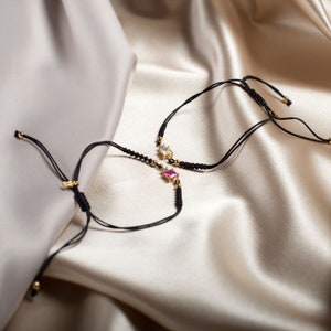 May include: Two adjustable black string bracelets with gold star charms. One charm has a pink gemstone and the other has a clear gemstone.