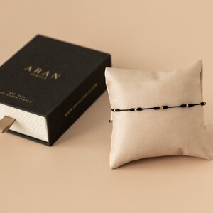 Puede incluir: Un brazalete de cuerda negra con cuentas doradas sobre un cojín beige. El brazalete se muestra en una caja negra con la palabra "ARAN" impresa en la parte superior.