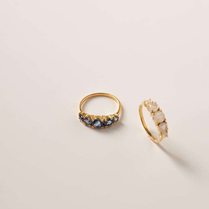 May include: Two gold rings with gemstones. The first ring has five blue gemstones set in a row. The second ring has five white gemstones set in a row.