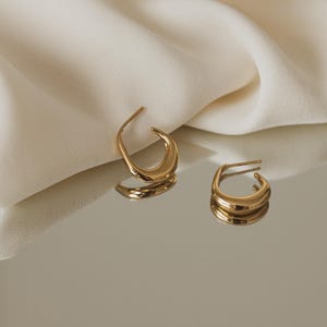 May include: A pair of gold hoop earrings with a curved, rounded shape. The earrings are on a white surface.